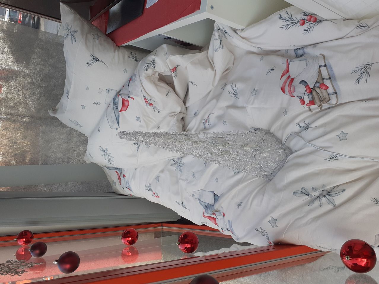 Eine silberne Weihnachtsbaumdekoration liegt auf einem weißen Bett mit weihnachtlich bedruckten Kopfkissen und einer Weihnachtsfigur. Darunter ist ein roter Glastisch mit Reflexionen roter Weihnachtskugeln.