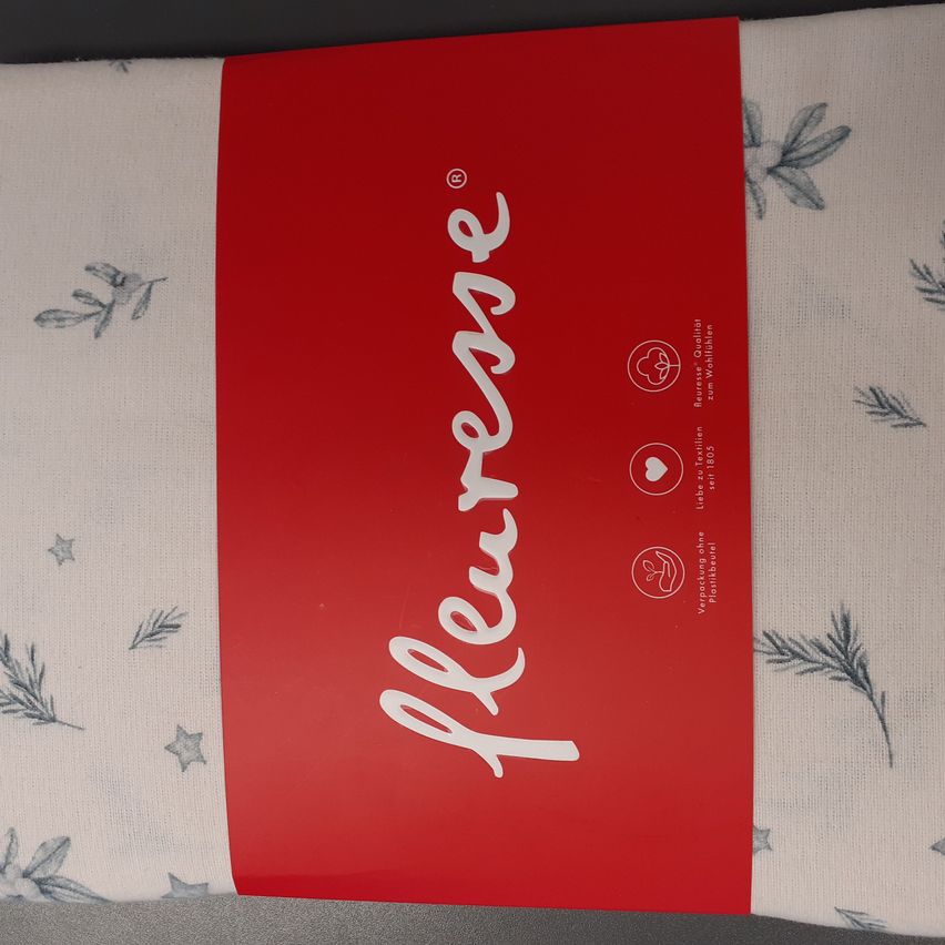 Ein rotes Stofflabel mit weißer Schrift liest sich 'Kleresse'. Der Stoff hat einen weißen Hintergrund mit grauen Blatt- und Sternenmustern. Ein Logo und Text befinden sich auf der rechten Seite des Labels.