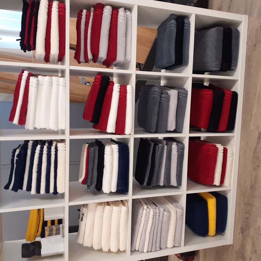 Ein weißer Regalschrank mit mehreren Fächern, jedes mit ordentlich gefalteten Handtüchern in verschiedenen Farben gefüllt, darunter Rot, Weiß, Grau und Blau. Die Handtücher sind in Reihen und Spalten angeordnet und schaffen eine ordentliche Präsentation.