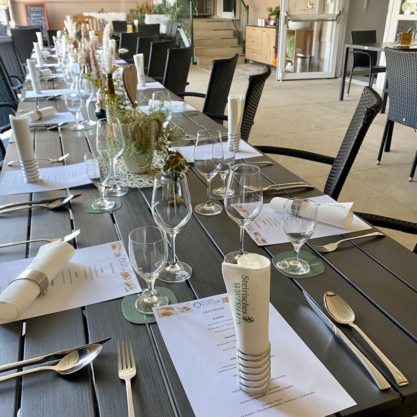 A long dining table set for a meal, with wine glasses, silverware, and a menu. The table is decorated with plants and paper napkins.