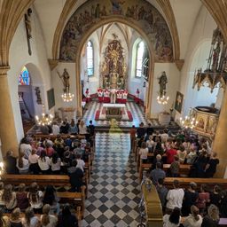 Ein Blick von oben auf das Innere einer Kirche mit Altar und Geistlichen vorne. Die Leute sitzen auf Bänken auf beiden Seiten. Die Kirche ist mit Kronleuchtern, Statuen und Gemälden geschmückt.