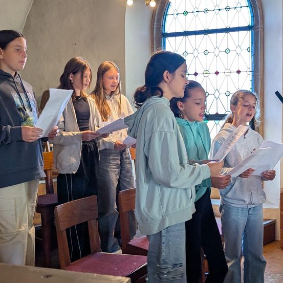 Eine Gruppe junger Mädchen steht in einer Kirche und hält Papiere in der Hand. Sie scheinen ein Lied zu singen oder eine Aufführung zu proben. Hinter ihnen stehen Holzstühle und Kirchenbänke.