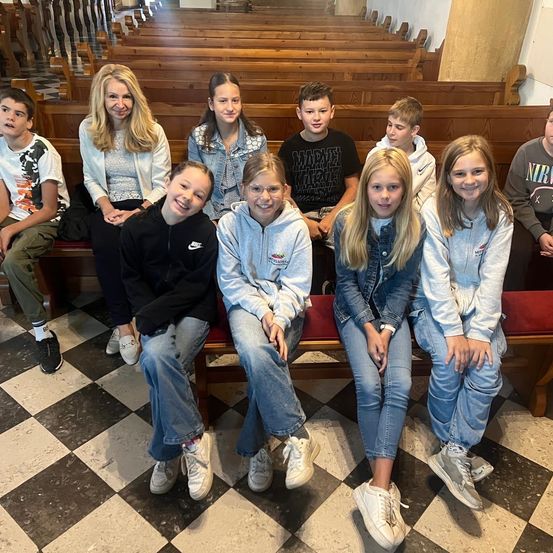 Eine Gruppe von Kindern und Erwachsenen, die auf einer Kirchenbank sitzen und für ein Foto lächeln. Die Bank ist rot und der Boden ist kariert. Die Kinder tragen Freizeitkleidung.