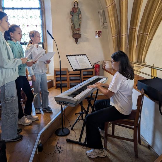 Eine Gruppe junger Mädchen steht in einer Kirche, singt und liest von einem Notenständer. Eine Frau spielt eine Tastatur und sitzt auf einem Stuhl. Ein Feuerlöscher und eine Statue sind an der Wand befestigt.