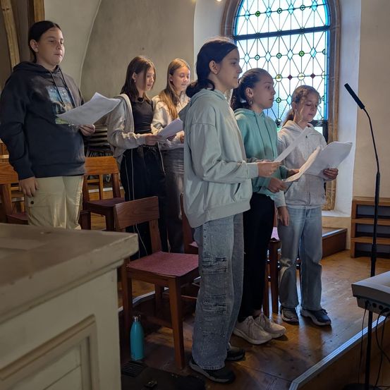 Eine Gruppe von Mädchen steht in einer Kirche, hält Papiere in der Hand und singt wahrscheinlich. Hinter ihnen sorgt ein Buntglasfenster für Licht.