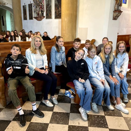 Eine Gruppe von Kindern und einer Frau posiert in einer Kirche, auf hölzernen Bänken sitzend. Die Kinder sind lässig gekleidet, und die Frau lächelt in die Kamera. Das Kircheninnere zeichnet sich durch religiöse Kunstwerke und einen gemusterten Boden aus.
