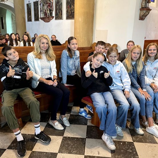 Eine Frau und mehrere Kinder sitzen gemeinsam auf einer Kirchenbank und posieren für ein Foto mit erhobenen Daumen.