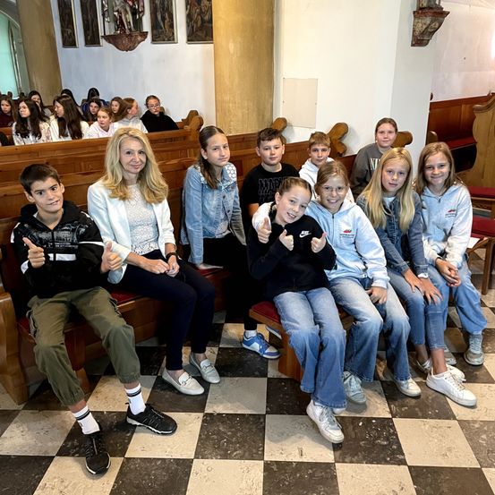Eine Gruppe von Kindern und einer erwachsenen Frau sitzen auf Kirchenbänken. Sie lächeln und zeigen Daumen hoch. Der Boden ist kariert.
