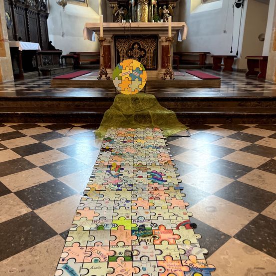 In einer Kirche führt eine lange Reihe von Puzzleteilen zu einem großen Puzzle auf dem Altar. Die Teile sind farbenfroh und haben Zeichnungen und Schriften. Der Altar hat Kerzen und eine Statue.