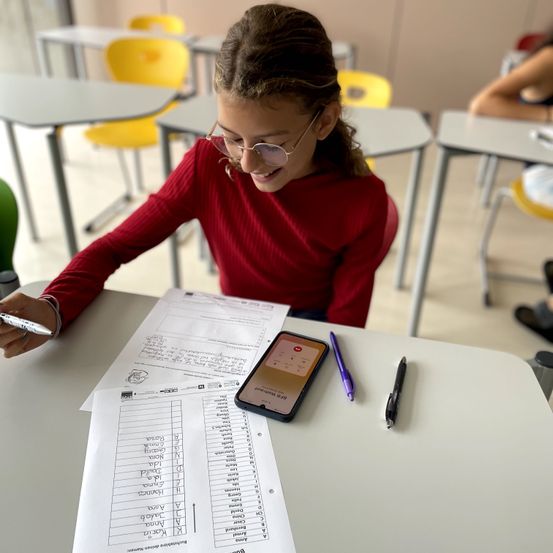Ein junges Mädchen sitzt in einem Klassenzimmer an einem Schreibtisch und hält einen Stift in der Hand. Sie lächelt. Auf dem Tisch liegen Papiere, ein Handy und Stifte. Andere Schüler sitzen im Hintergrund an ihren Schreibtischen.