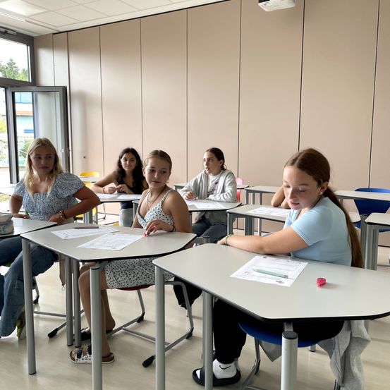 Fünf Mädchen sitzen in einem Klassenzimmer. Sie arbeiten mit Stiften an Papieren. Ein Mädchen schaut auf ihr Papier, während ein anderes Mädchen lächelt.
