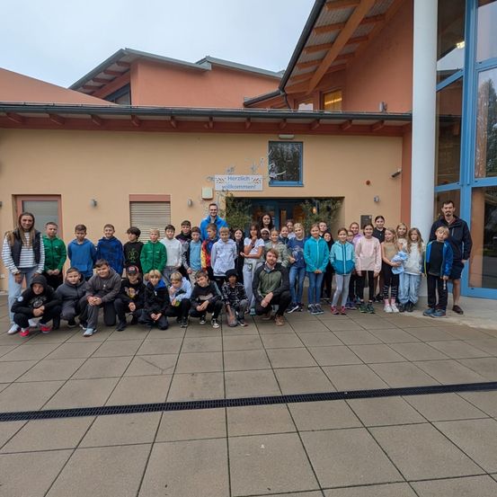 Eine Gruppe von Kindern und Erwachsenen posiert für ein Foto vor einem Gebäude. Das Gebäude hat Glasfenster und ein Schild, das Herzlich Willkommen steht.