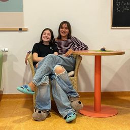 Eine Frau und ein junges Mädchen sitzen auf einem Stuhl in einem Raum mit einem runden Tisch. Sie lächeln beide und tragen Hausschuhe. Eine Tafel hängt an der Wand.