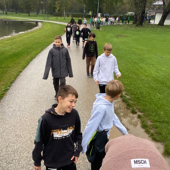 Kinder laufen auf einem gepflasterten Weg in einem Park mit einem Teich, einige tragen Hoodies und Turnschuhe. Ein Junge mit einer Mütze ist im Vordergrund zu sehen.