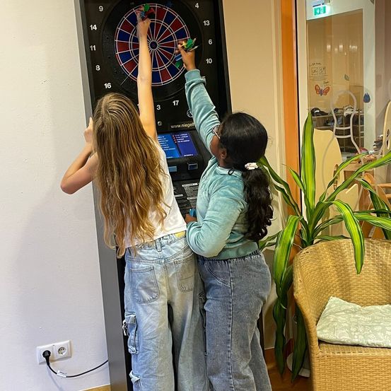 Zwei Mädchen stehen vor einem Dartboard. Ein Mädchen hält einen Dart und zeigt auf das Brett, während das andere Mädchen ebenfalls einen Dart hält.