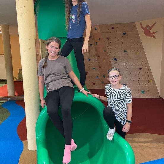 Drei Mädchen spielen in einem Indoor-Spielplatz. Ein Mädchen sitzt auf einem grünen Rutschvergnügen, ein anderes steht auf dem Rutschvergnügen und das dritte steht neben dem Rutschvergnügen. Sie alle lächeln. Der Rutschvergnügen befindet sich in der Nähe einer Kletterwand.
