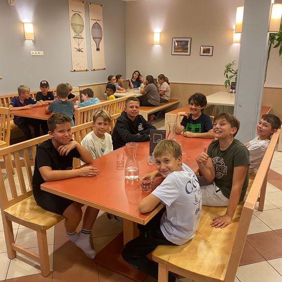 Eine Gruppe junger Jungen sitzt in einem Restaurant an Tischen, lächelt und posiert für ein Foto. Der Raum hat weiße Wände, einige Bilder und Lampen. Einige Leute sitzen an anderen Tischen.