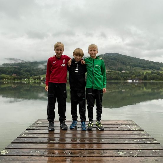 Drei Jungen stehen auf einem Steg am See mit Bergen im Hintergrund. Einer trägt ein rotes Shirt, ein anderer ein schwarzes Jacket und der dritte ein grünes Jacket.