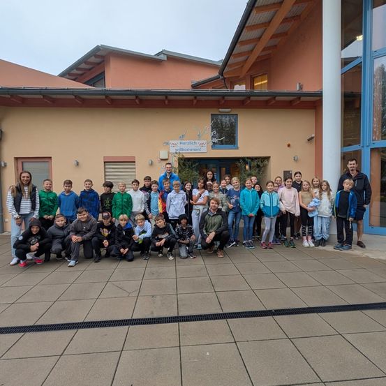 Eine Gruppe von Schülern und Lehrern posiert für ein Foto vor einem Gebäude mit einem Schild, das Herzlich willkommen! bedeutet.