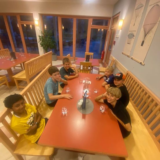 Eine Gruppe von Kindern und Jugendlichen sitzt um einen roten Tisch in einem Essbereich und posiert für ein Foto. Sie sind von Stühlen und einer Pflanze umgeben.