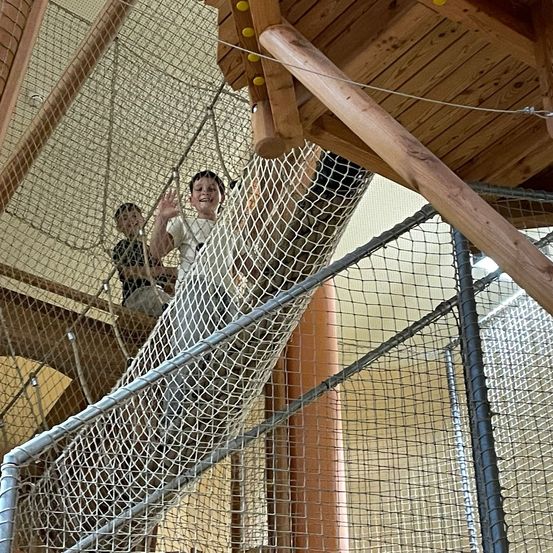 Zwei Kinder klettern auf einer Netzleiter in einer Spielstruktur. Eines winkt. Die Struktur hat Holzbalken und Netzgitter.