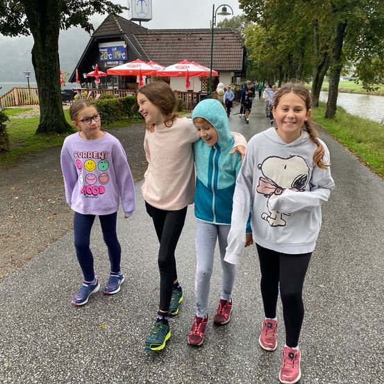 Vier Mädchen gehen auf einem Pfad neben einem Fluss, lächeln und posieren für ein Foto. Hinter ihnen steht ein kleines Gebäude mit einem Wetterschild, und mehrere Menschen gehen in der Ferne.