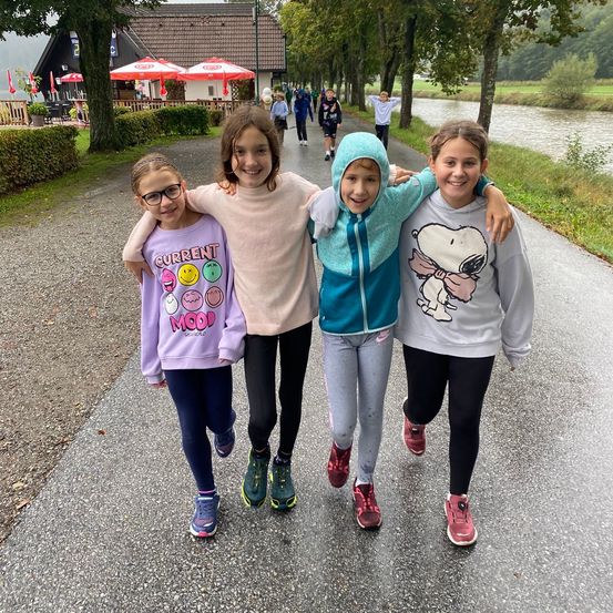 Vier junge Mädchen gehen gemeinsam auf einem Pfad neben einem Fluss und lächeln für die Kamera. Sie sind in verschiedenen Farben und Stilen gekleidet, eine trägt eine Brille. Hinter ihnen gehen weitere Kinder und ein Gebäude mit roten Schirmen.