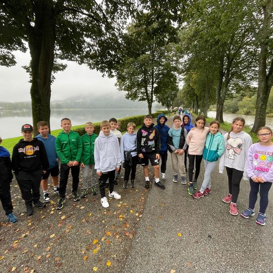 Eine Gruppe von Kindern und Jugendlichen posiert für ein Foto auf einem Weg in der Nähe eines Sees, mit Bäumen und Büschen im Hintergrund.