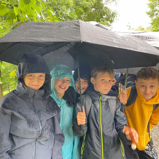 Fünf Kinder lächeln und posieren für ein Foto, alle in Regenmänteln und mit Regenschirmen unter dem Regen.