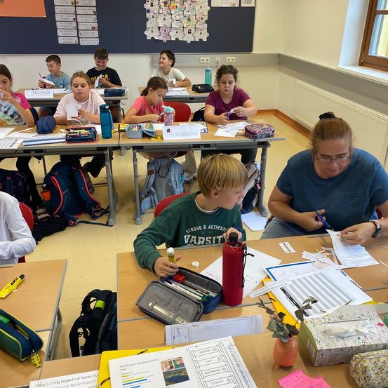 In einem Klassenzimmer sitzen Schüler an Tischen und arbeiten an Aufgaben. Eine Lehrerin hilft einem Jungen vorne, während andere sich auf ihre Aufgaben konzentrieren. Die Tische sind mit Papieren, Stiften und anderen Schulsachen überladen.