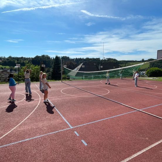 Eine Gruppe von Menschen spielt Volleyball auf einem Freiluftplatz. Sie tragen Freizeitkleidung und Turnschuhe. Der Platz ist von einem Zaun mit Netz umgeben. In der Ferne befinden sich Bäume und Häuser.