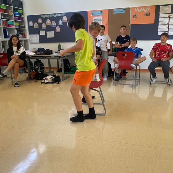 In einem Klassenzimmer übt ein Junge in einem gelben T-Shirt und orangefarbenen Shorts Tanz. Mehrere Kinder sitzen in roten Stühlen und beobachten. Eine Frau in einem schwarzen Shirt sitzt in einem Stuhl. Rucksäcke und Gegenstände sind auf einem Tisch.