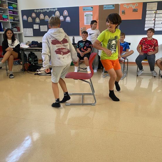 Zwei Jungen tanzen in einem Klassenzimmer. Einer trägt eine Kapuzenjacke, der andere ein grünes T-Shirt und orange Shorts. Hinter ihnen sitzen mehrere Kinder auf Stühlen und beobachten. Eine Frau sitzt auf einem Stuhl links, und hinter ihr steht ein Regal voller Bücher und Gegenstände.