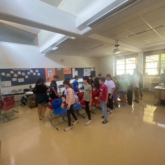 Eine Gruppe von Kindern und Erwachsenen in einem Klassenzimmer mit einer Lehrerin, mehreren Stühlen und Ankündigungstafeln an der Wand.