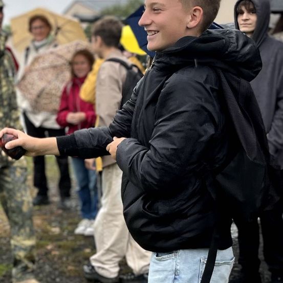 Ein Junge mit Rucksack hält einen Stein und lächelt andere in einer Gruppe an, einige halten Regenschirme.
