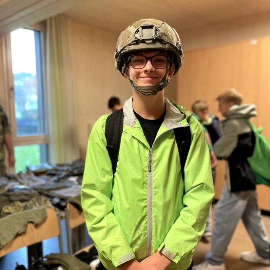 Ein Junge in einer grünen Jacke und einem Militärhelm steht in einem Raum mit Tischen voller Militärkleidung. Andere Jungen sind im Hintergrund zu sehen, einer trägt einen Rucksack.