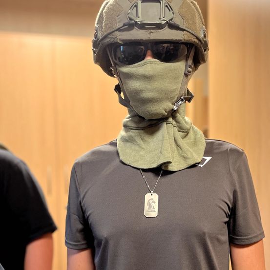 Eine Person in Camouflage-Ausrüstung steht vor einer Holzwand, trägt einen Helm, Sonnenbrille und einen grünen Schal über dem Gesicht. Sie trägt ein graues T-Shirt und eine Hundemarkenkette.