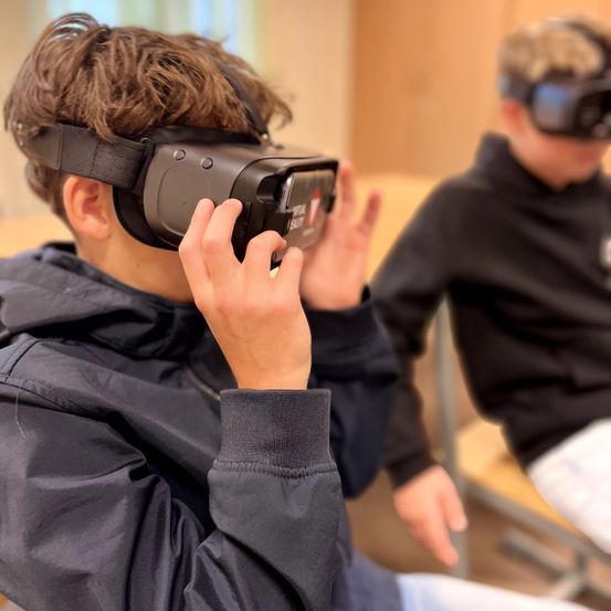 Zwei Jungen sitzen drinnen und tragen VR-Headsets, einer hält einen Controller. Der Junge im Vordergrund hat einen ernsten Ausdruck.