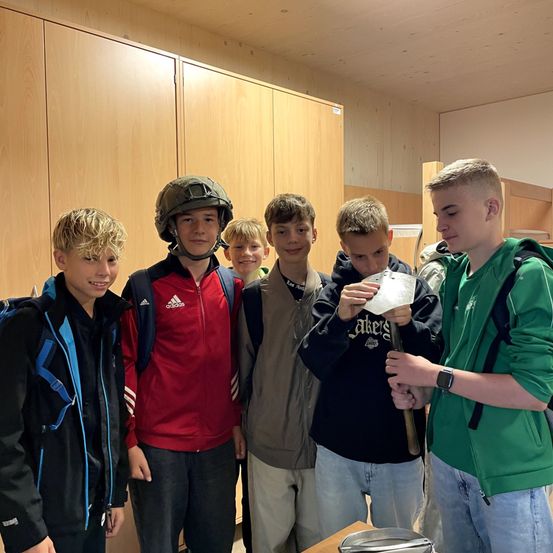 Eine Gruppe junger Jungen steht in einem Raum zusammen. Ein Junge hält ein Stück Papier und ein anderer Junge trägt einen Helm.