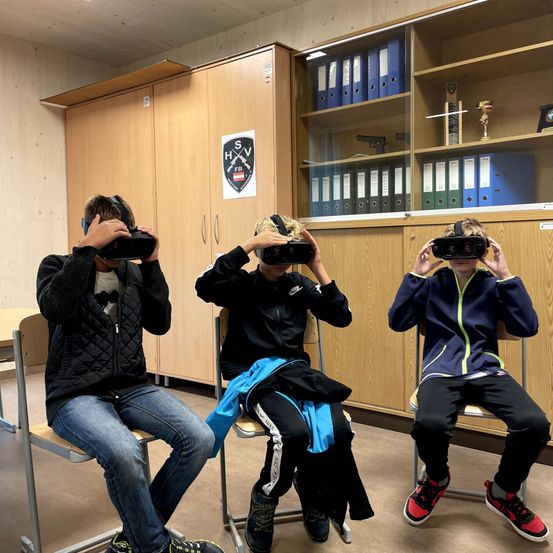 Drei Kinder sitzen auf Stühlen, tragen VR-Headsets und schauen in eine virtuelle Umgebung. Dahinter steht ein Schrank mit Ordnern und Trophäen. Eine weiße Tafel mit schwarzen Buchstaben ist an der Wand angebracht.