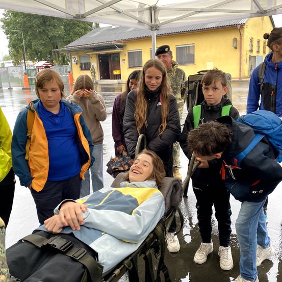 Bei Regen lächelt ein Mädchen auf einer Trage Schulkinder unter einem Zelt an, während ein Mann in Uniform hinter ihr steht.