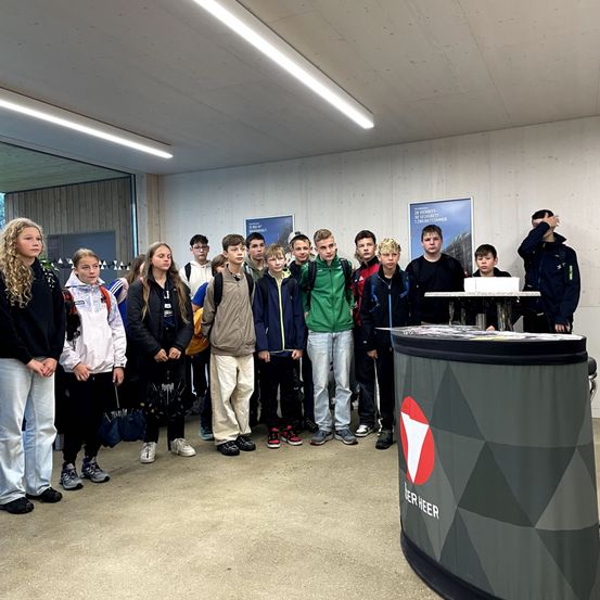 Eine Gruppe von Schülern steht in einem modernen, gut beleuchteten Raum, mit zwei Erwachsenen vorne. Es gibt einen Stand mit einem roten und weißen Logo und Informationsposter an der Wand.