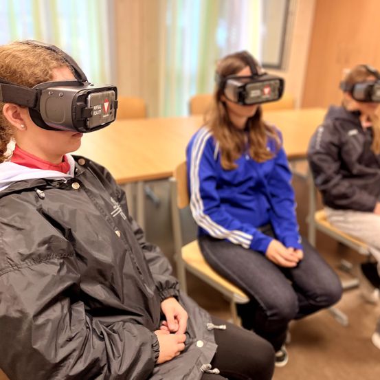 Drei Personen tragen Virtual-Reality-Headsets in einem Raum. Die Person vorne trägt eine schwarze Jacke. Die Person dahinter trägt eine blaue Jacke. Die Person rechts trägt eine schwarze Jacke.