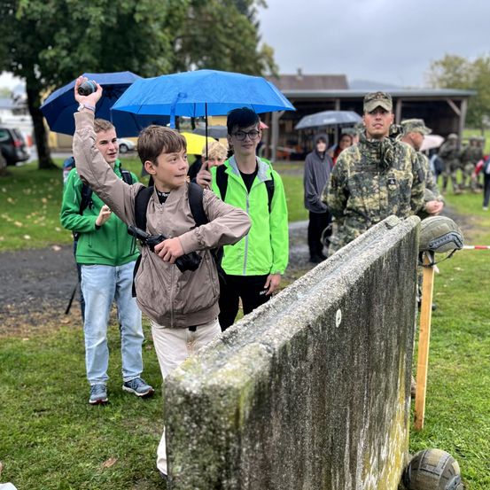 Ein Junge hält einen Ball und wirft ihn in einem Park. Ein anderer Junge steht hinter ihm, und zwei weitere stehen neben ihm. Einige Leute stehen hinter ihnen.