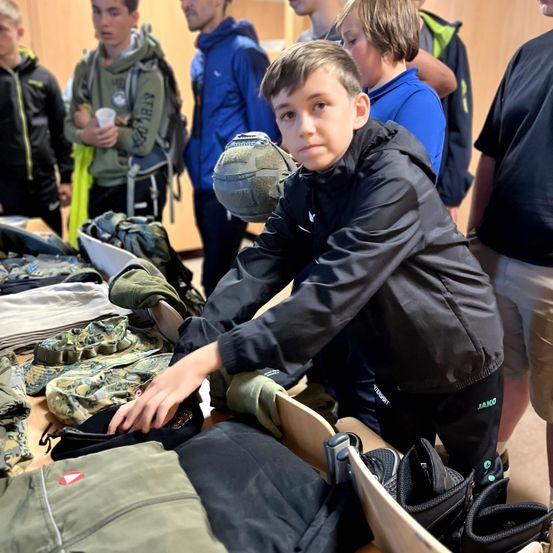 Ein Junge in einer schwarzen Jacke untersucht verschiedene Tarnkleidungsstücke und Ausrüstungsgegenstände, die auf einem Tisch in einem Raum mit anderen Kindern ausgelegt sind.
