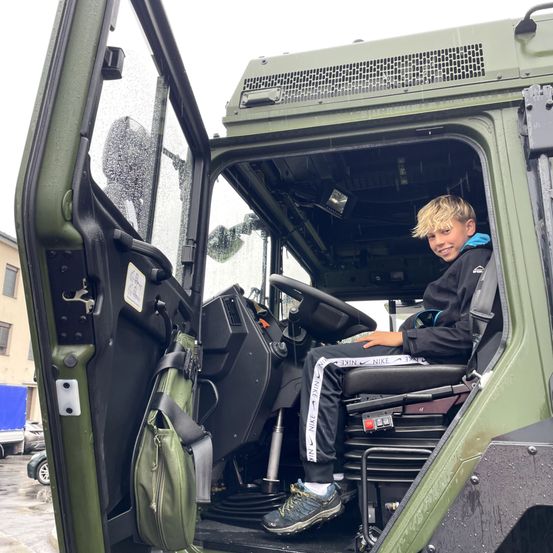 Ein junger Junge sitzt lächelnd in einem grünen Militärfahrzeug. Er trägt eine blaue Kapuzenjacke, schwarze Hosen und Turnschuhe. Eine grüne Tasche hängt an der Tür, und das Armaturenbrett ist sichtbar.