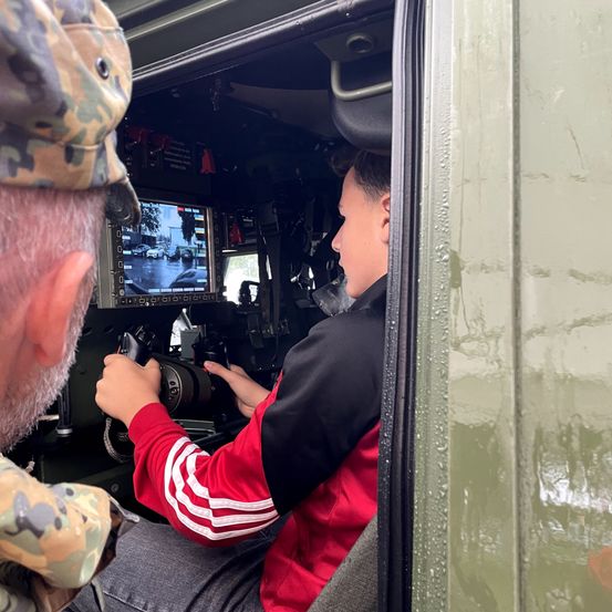 Ein Soldat beobachtet, wie ein junger Mann ein Militärfahrzeug fährt, wobei der Monitorbildschirm die Aussicht von draußen zeigt.