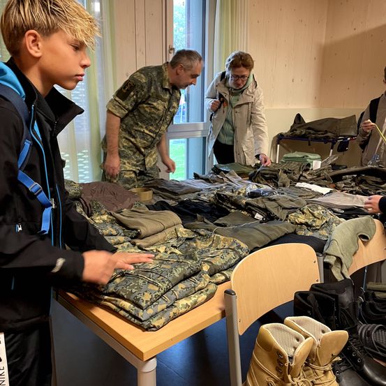 Ein Junge und ein Mann in Uniform inspizieren Militärkleidung auf einem Tisch mit Stiefeln in der Nähe.