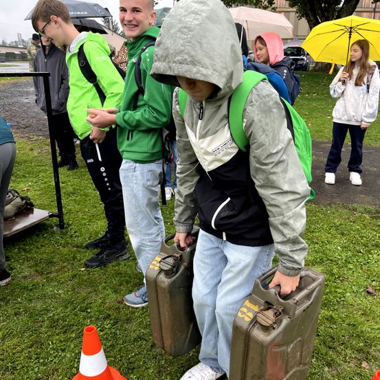 Eine Gruppe junger Leute steht auf einem Rasenfeld, einer von ihnen hält zwei große Kanister. Sie sind in verschiedene Jacken gekleidet und einige halten Regenschirme. Im Hintergrund sind weitere Menschen, Autos und ein Baum zu sehen.