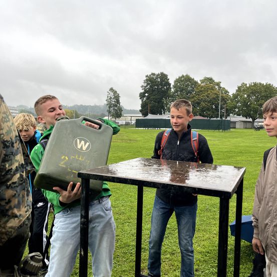 Mehrere Jungen stehen im Freien. Ein Junge hält einen Metallbehälter, während ein anderer ihn ansieht. Sie sind draußen auf einem Feld.